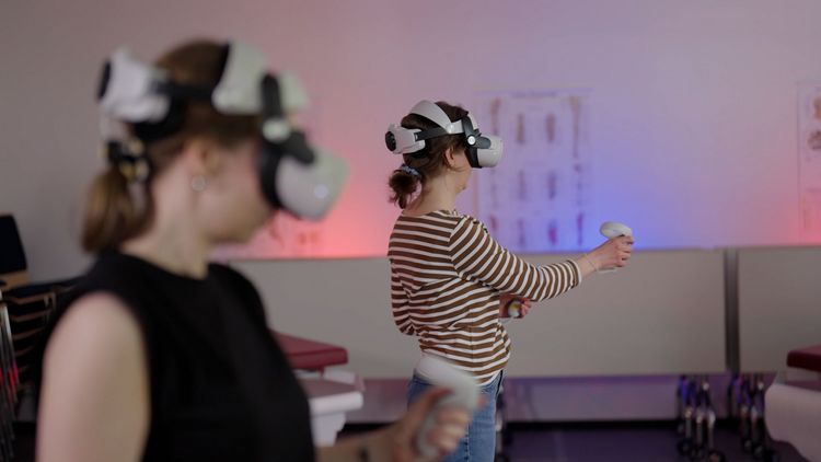 Zwei Personen mit VR-Brille über den Augen stehen in einem Seminarraum und hantieren mit Controllern. 