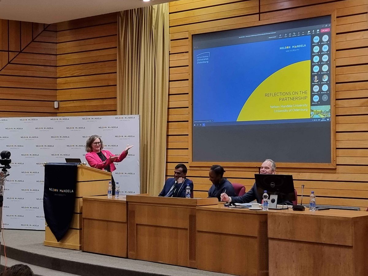 Delegationsbesuch an der NMU Eine Frau spricht hinter einem Pult, während andere Personen ihr zuhören. Im Hintergrund ist eine Fotowand mit dem Logo der Nelson Mandela University.