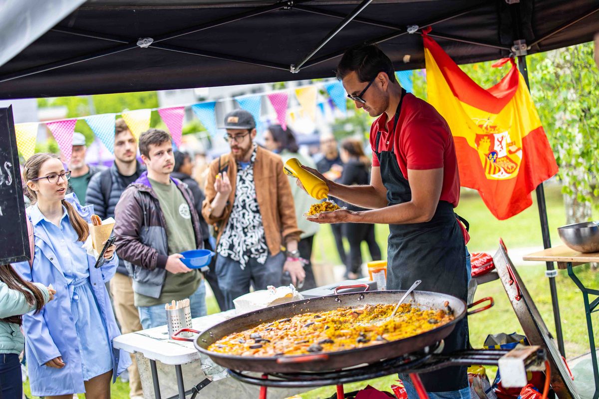 Ein mit spanischer Flagge geschmückter Essensstand. Eine Person bereitet einen Teller vor, der aus einer riesigen Paella-Pfanne kommt. Wartende Interessierte im Hintergrund.