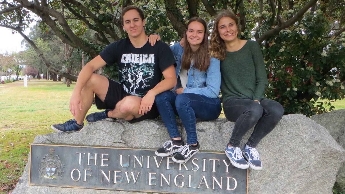 Das Bild zeigt drei lächelnde junge Menschen, die auf einem Felsen sitzen. Vor ihnen befindet sich eine bronzene Plakette mit dem Wappen und dem Schriftzug "The University of New England". Im Hintergrund erstreckt sich eine grüne Rasenfläche mit Bäumen und Gebäuden der Universität. Der Himmel ist leicht bewölkt und die Sonne scheint.