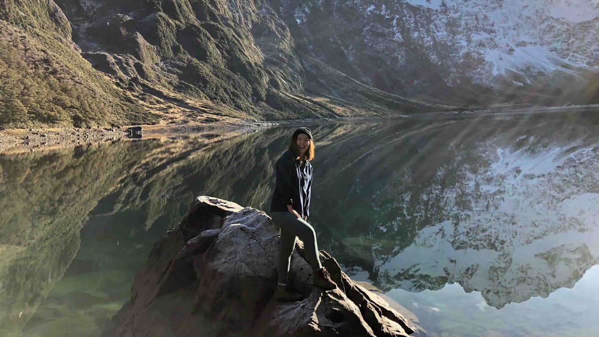 Das Bild zeigt eine Frau, die auf einem Felsen am Ufer eines Sees steht. Hinter ihr erheben sich hohe, schneebedeckte Berge, die sich im ruhigen Wasser des Sees spiegeln. Die Frau trägt eine Mütze und eine Jacke und blickt in die Kamera.