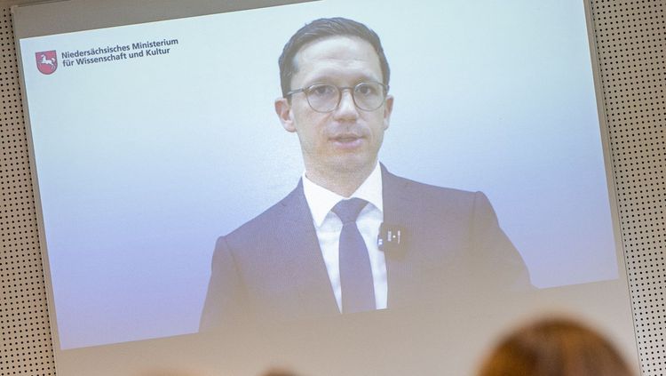 Das Bild zeigt Minister Falko Mohrs. Er hält eine Videoansprache, die per Beamer auf eine Wand projiziert wird. Der Minister schaut ernst und gestikuliert leicht während seines Vortrages.