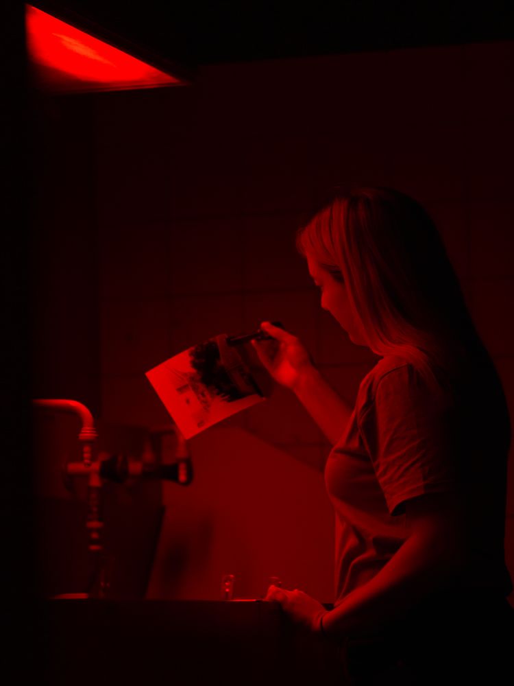 Analoges Arbeiten in der Dunkelkammer — Anna Siebert — Im Rahmen meiner Bachelorarbeit möchte ich mich mit dem Wiederaufkommen der Analogfotografie im digitalen Zeitalter des 21. Jahrhunderts beschäftigen. Auf dieses Thema gestoßen bin ich über mein Hobby, vorwiegend digital und gelegentlich analog zu fotografieren. Meine Fotografie zeigt eine Studentin beim Entwickeln eines analogen Filmes im Fotolabor. 