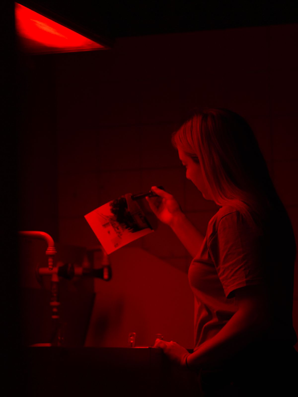 Analoges Arbeiten in der Dunkelkammer — Anna Siebert — Im Rahmen meiner Bachelorarbeit möchte ich mich mit dem Wiederaufkommen der Analogfotografie im digitalen Zeitalter des 21. Jahrhunderts beschäftigen. Auf dieses Thema gestoßen bin ich über mein Hobby, vorwiegend digital und gelegentlich analog zu fotografieren. Meine Fotografie zeigt eine Studentin beim Entwickeln eines analogen Filmes im Fotolabor. 