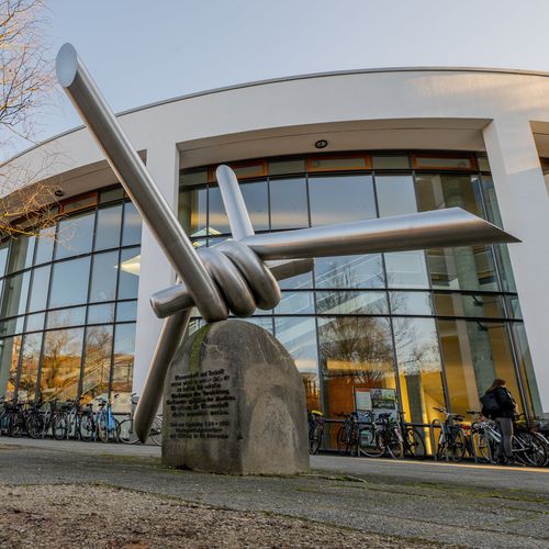 Das Bild zeigt das Mahnmal für Ossietzky vor dem Audimax. Es stellt einen Stacheldraht dar. Vorne zu erkennen ist die im Text erwähnte Inschrift. Im Hintergrund stehen Fahrräder, die Sonne scheint.
