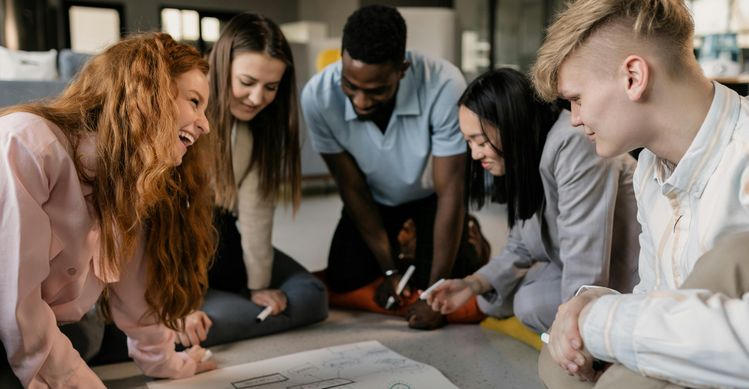 Interkulturelles Training Im Bild ist eine Gruppe von fünf Personen um einen Tisch versammelt, die an einer gemeinsamen Aktivität beteiligt sind. Sie lächeln und scheinen gemeinsam an einem Projekt zu arbeiten, was auf eine positive und inklusive Atmosphäre hinweist.