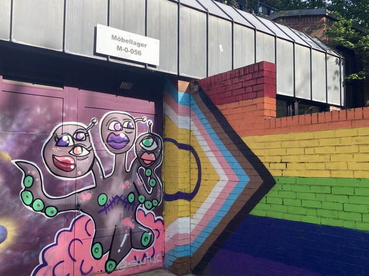 The picture is a photo of the entrance door to the furniture warehouse. There is purple graffiti on the door and on the right you can see a wall with the rainbow flag of the LGBTQI+ movement painted on it.