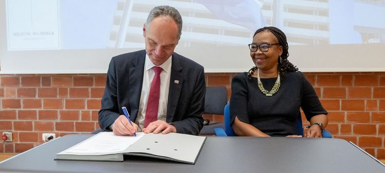 25-jähriges-Kooperationsjubiläum Das Bild zeigt zwei Personen, Vice-Chancellor Sibongile Muthwa (NMU) und Präsident Ralph Bruder (UOL), die an einem Tisch sitzen und ein Dokument unterzeichnen. Der Präsident der Universität Oldenburg schreibt mit einem Stift auf das Dokument. Die Vice-Chancellor der Nelson Mandela University lächelt und blickt auf das Dokument. Im Hintergrund ist ein Banner mit der Aufschrift "University of Oldenburg | President" und ein Bild des Universitätsgebäudes der NMU zu sehen. Das Bild dokumentiert die Unterzeichnung einer Vereinbarung im Rahmen des 25-jährigen Kooperationsjubiläums zwischen den beiden Universitäten.
