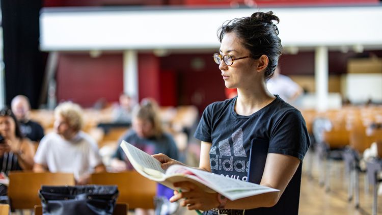 Die Person im Vordergrund mit Textbuch, dahinter sind unscharf die Stuhlreihen der Aula zu sehen, wo auch einige Leute sitzen. 