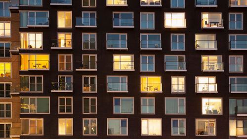The picture shows an exterior view of a residential building at dusk. Lights are switched on in many of the rooms in the multi-storey building.