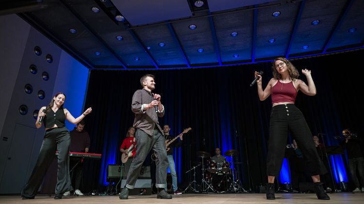 The picture shows the band SWAY at a performance. Three band members play wind instruments, one drums, two play guitar, one keyboard. Three others sing and dance.