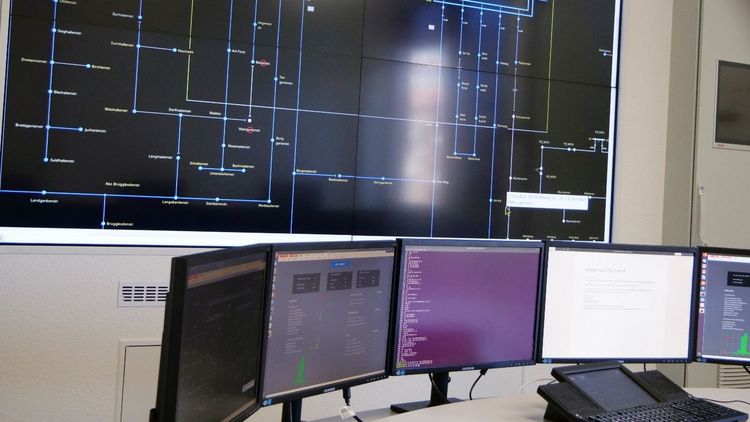 Ein großser Bildschirm mit einem Schaltbild an der Wand, darunter sind mehrere große Computerbildschirme nebeneinander aufgereiht, ähnlich wie in einem Kontrollzentrim