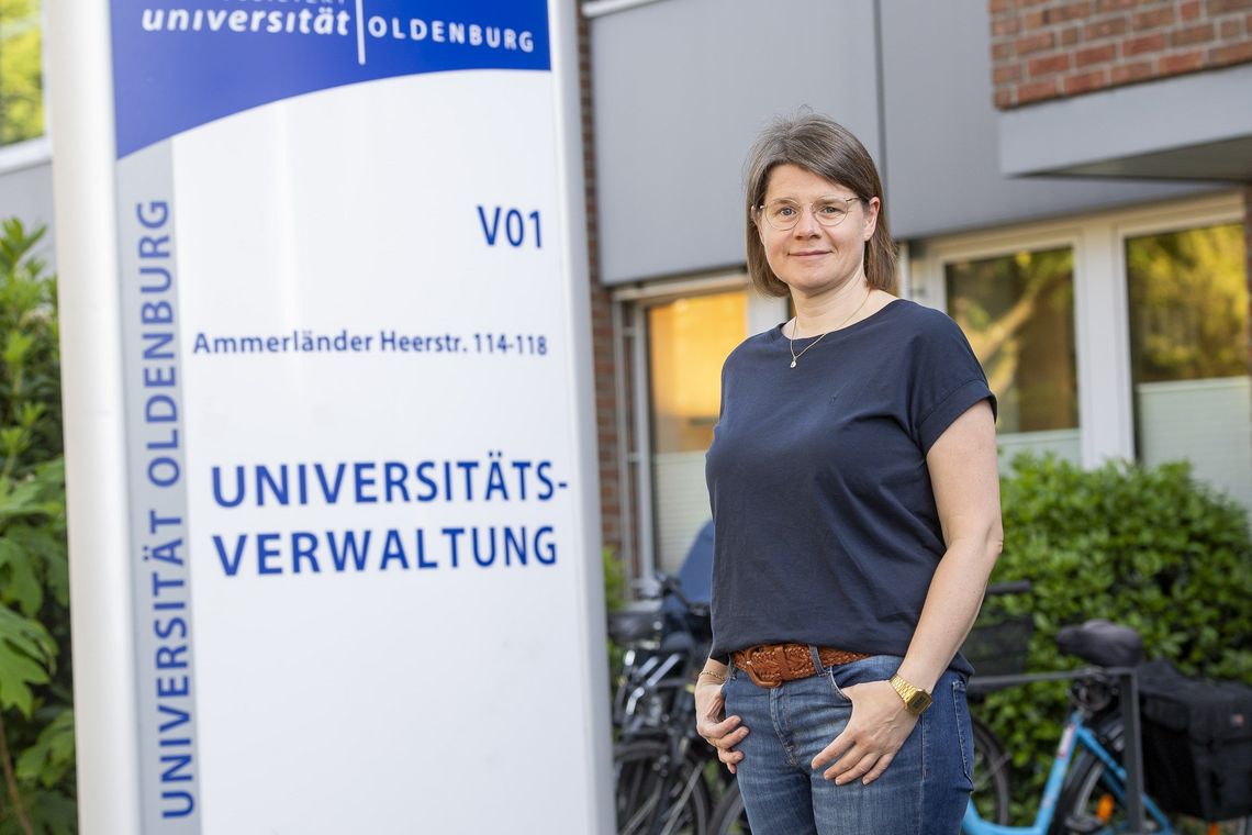 Imke Hansjürgens steht vor dem Eingang zu V01. Hinter ihr ist das große Schild zu erkennen, auf dem "Universitätsverwaltung" steht. Sie schaut in die Kamera und hat die Daumen in den Hosentaschen.
