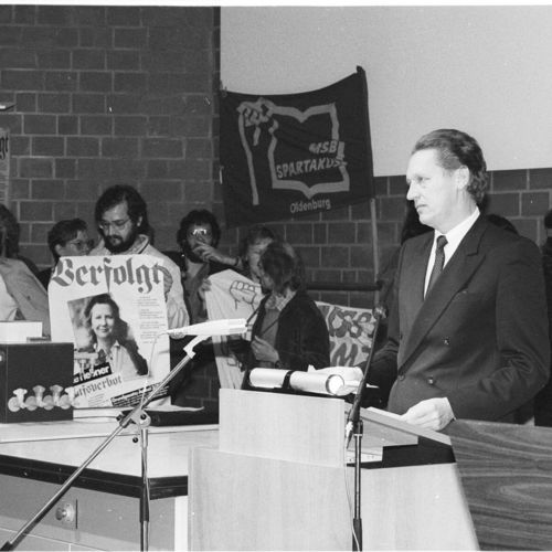 Das Bild zeigt den damaligen niedersächsischen Minister für Wissenschaft und Kunst Dr. Johann-Tönjes Cassens. Er ist umringt von Studenten, während er eine Rede halten will. Sie unterbrechen ihn und halten Plakate von Menschen hoch, die Aufgrund der Berufsverbote ihre Arbeitsstelle verloren haben. Der Minister schaut indigniert.