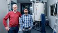 Two men stand in front of a large technical device. Numerous metal hoses and pipes protrude from the top and bottom of a barrel-shaped device. These are the cooling chambers inside which the measurement takes place. 