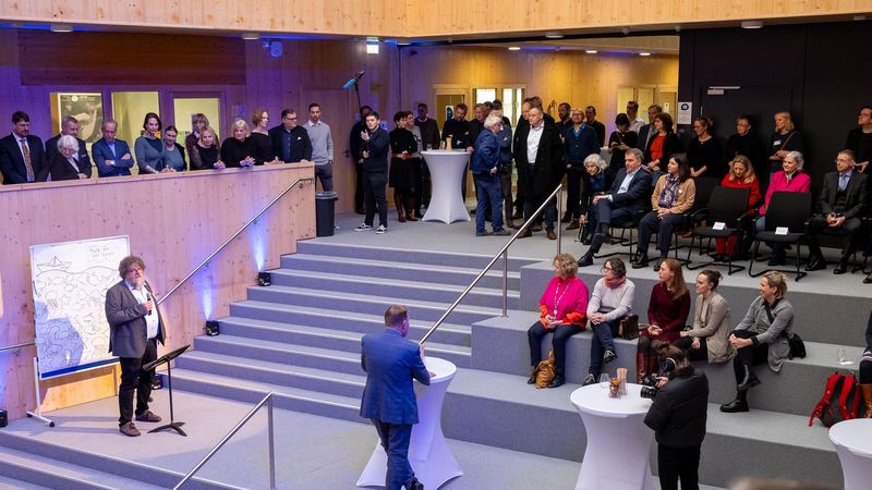 Blick in den Innenraum des HIFMB, einige Personen stehen an Stehtischen, andere Sitzen auf den Stufen, die meisten stehen auf einer Art Gallerie. Ein Mann steht etwas getrennt von den Gästen auf einem Absatz hinter einem Mikrofon.