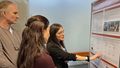 Two young women and an older man look at a scientific poster together and discuss it.