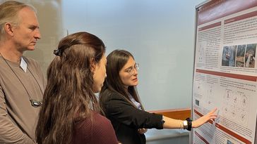 Zwei junge Frauen und ein älterer Mann betrachten gemeinsam ein wissenschaftliches Poster und diskutieren.