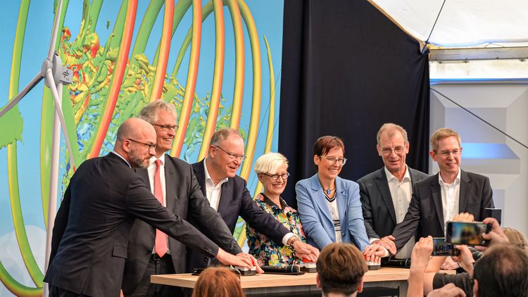 Die sechs Personen stehen hinter einem Tisch, drücken auf mehrere große, rote Knöpfe zur Eröffnung. Im Hintergrund ist die Simulation eines Windrads mit turbulenten Luftströmungen zu sehen.