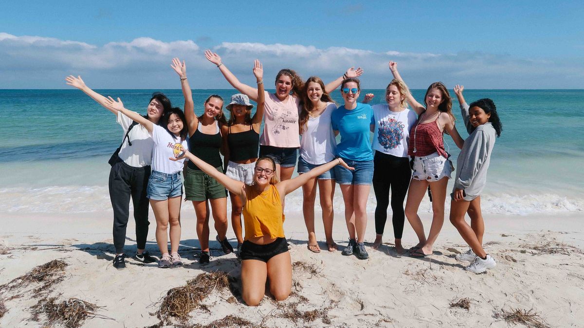 Das Bild zeigt eine Gruppe von jungen Frauen, die am Strand mit erhobenen Armen posieren. Sie stehen auf weißem Sand vor türkisfarbenem Meer und einem blauen Himmel mit vereinzelten Wolken. Die Frauen lächeln und wirken fröhlich und entspannt. Die Szene vermittelt ein Gefühl von Gemeinschaft und Spaß.