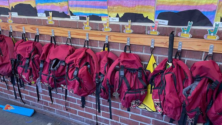 Praktikum im Ausland (Schule) Ein Klassenraum oder Flur in einer Schule mit kindlichen Malereien von Bergen und einem Sonnenuntergang. Darunter hängen rote Schultaschen mit Lamas und Kindernamen.