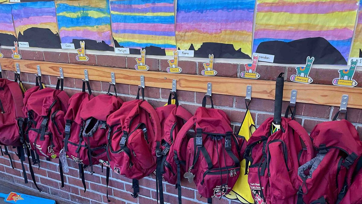 Praktikum im Ausland (Schule) Ein Klassenraum oder Flur in einer Schule mit kindlichen Malereien von Bergen und einem Sonnenuntergang. Darunter hängen rote Schultaschen mit Lamas und Kindernamen.