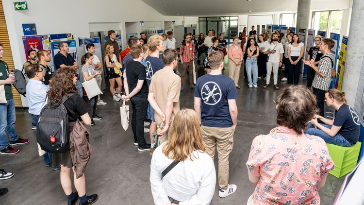 Numerous posters are set up in the foyer of the lecture theatre centre. A large number of people stand in a circle and listen to a student standing in front of his poster and explaining his research.