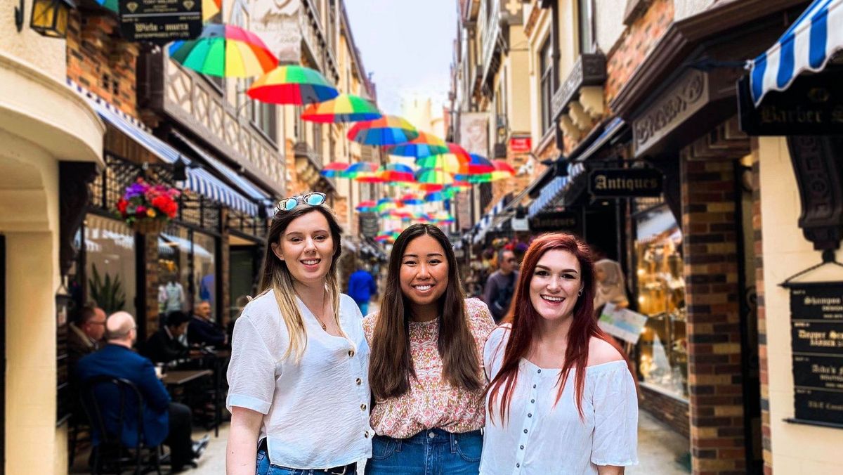 Das Bild zeigt drei junge Frauen, die in einer relativ engen Gasse zwischen historischen Gebäuden stehen. Über der Gasse sind bunte Regenschirme gespannt, die einen farbenfrohen Kontrast zu den alten Steinfassaden bilden. Die Frauen lächeln in die Kamera und wirken fröhlich. Im Hintergrund sind Geschäfte und Restaurants zu sehen. Die Szene vermittelt ein Gefühl von urbanem Charme und Lebendigkeit.