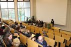 Auf dem Bild ist ein Hörsaal zu sehen, das Bild ist in Richtung Vortragender aufgenommen. Hinter dem Rednerpult steht eine Person, die Reihen sind gefüllt.