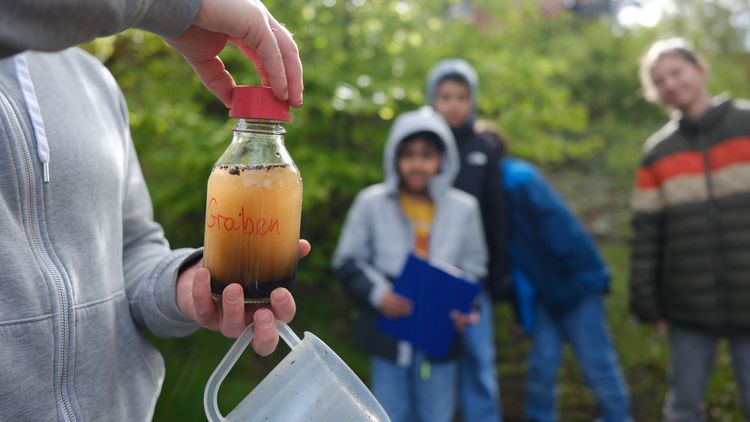 Einige Kinder schauen auf eine Flasche mit einer orangenen Flüssigkeit, auf der "Graben" steht.