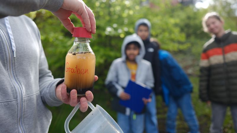 Einige Kinder schauen auf eine Flasche mit einer orangenen Flüssigkeit, auf der "Graben" steht.