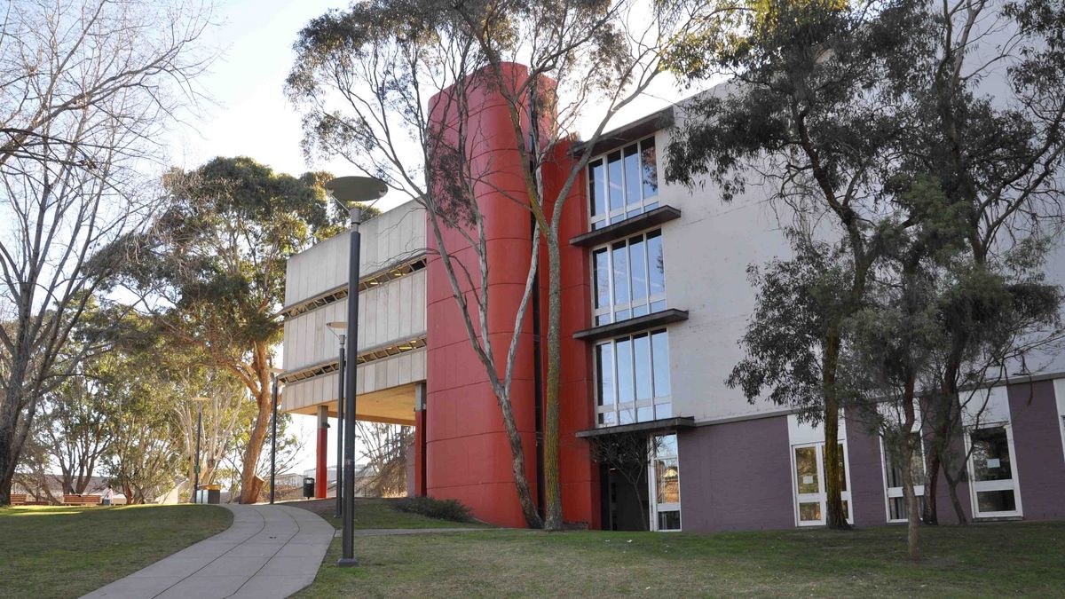 Das Bild zeigt ein markantes Gebäude auf dem Campus der University of New England. Das Gebäude zeichnet sich durch seine moderne Architektur aus, mit einer Kombination aus Beton, Glas und einem auffälligen roten Zylinder. Bäume umrahmen das Gebäude und ein Fußweg führt zum Eingang. Der Himmel ist blau und die Sonne scheint.