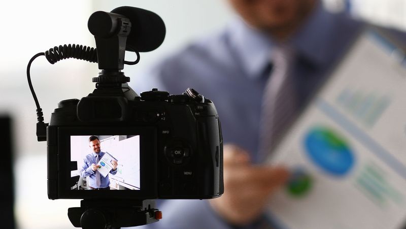 Videokamera im Vordergrund, auf dem Display ist ein Mann zu sehen, der ein Blatt Papier mit einigen Grafiken erklärt.