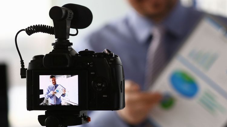Videokamera im Vordergrund, auf dem Display ist ein Mann zu sehen, der ein Blatt Papier mit einigen Grafiken erklärt.