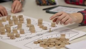 Das Bild zeigt das Spielfeld in Nahaufnahme. Hölzerne Chips liegen auf dem Feld. Sie stellen unterschiedlich hoch gewachsene Bäume dar. Eine Studentin zeigt mit Ihrer Hand auf einen der Bäume.