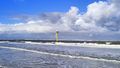The picture shows a yellow measuring pole standing in the North Sea on the beach off Spiekeroog. It is surrounded by water, waves are crashing on the beach. The sky is blue, some white and grey clouds can be seen.