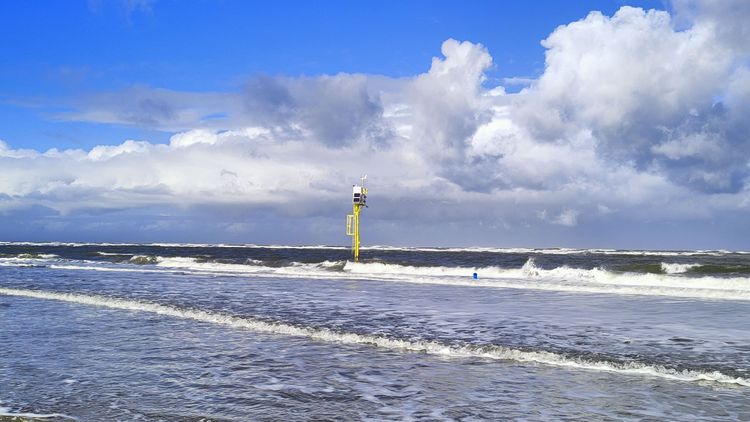 Das Bild zeigt einen gelben Messpfahl, der in der Nordsee am Strand vor Spiekeroog steht. Er ist umgeben von Wasser, Wellen branden am Strand an. Der Himmel ist blau, einige weiße und graue Wolken sind zu erkennen.