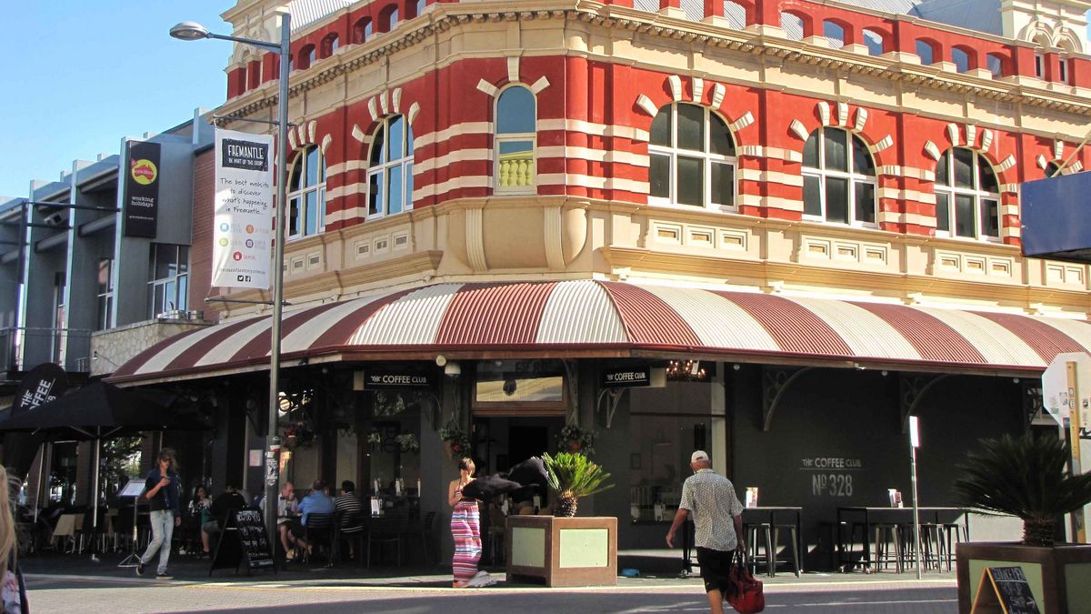 Das Bild zeigt das historische Fremantle Post Office in Fremantle, Westaustralien, mit seiner markanten rot-weiß gestreiften Fassade. Unter dem Gebäude befindet sich ein Café mit einer Markise und Tischen im Freien. Menschen gehen auf dem Bürgersteig vorbei. Das Gebäude ist ein Wahrzeichen der Stadt.