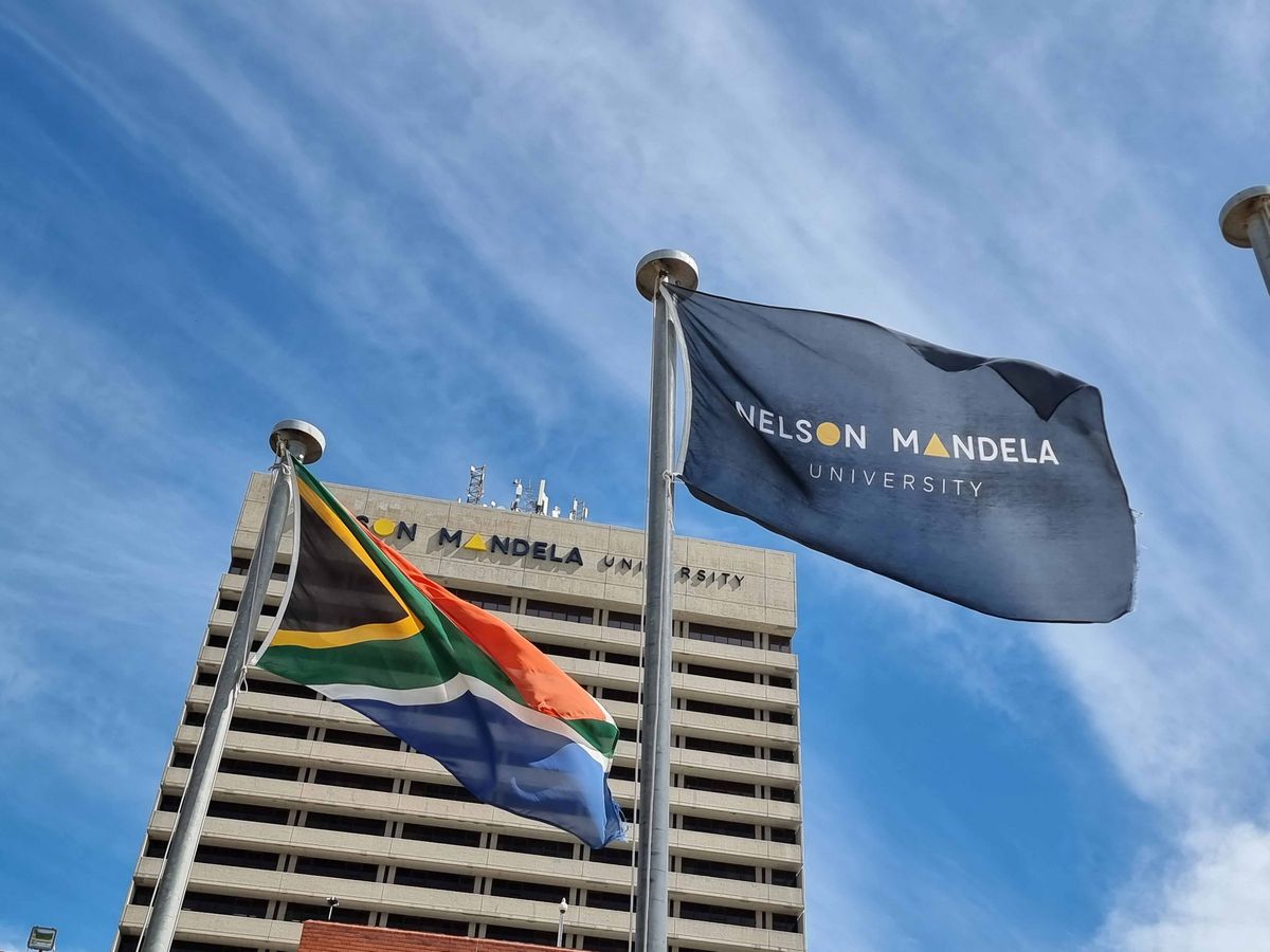 Nelson Mandela Unviersity Flaggen der Nelson Mandela University und Südafrikas wehen im Wind vor einem hohen Gebäude.