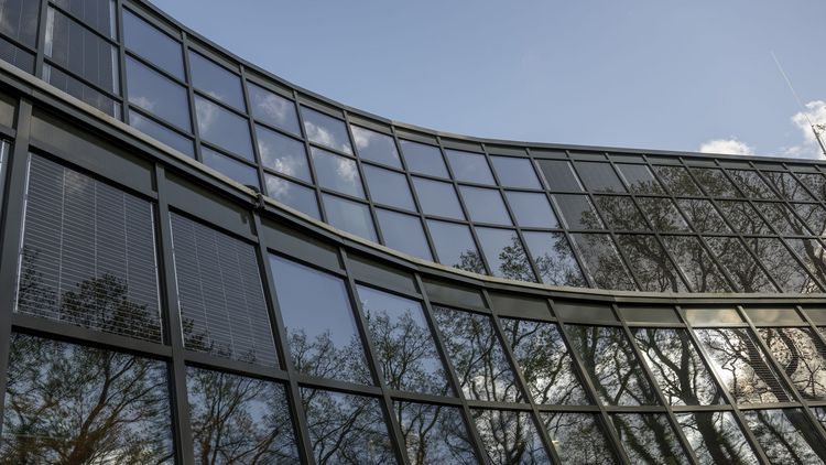 Auf den Scheiben der Glasfassade spiegeln sich der blaue Himmel, die Bäume und einige Wolken.. 