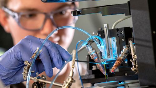 Eine Person mit Handschuhen nimmt Einstellungen am 3D-Drucker vor, aus dem Schläuche ragen. 