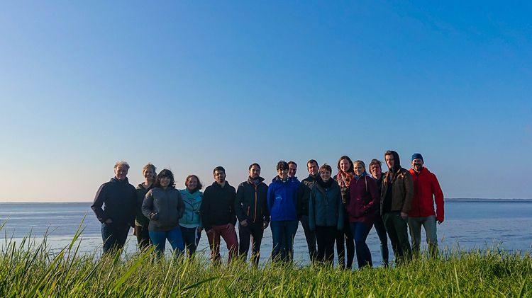 Gruppe aus 15 Personen steht auf einer Wiese, im Hintergrund das Meer und blauer Himmel. 