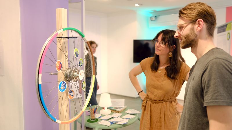 Anna Krämer zeigt einem Besucher des Stores einen Ausstellungsgegenstand in Form eines Fahrrad-Rades.