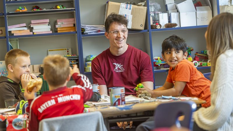 Ein Student, eine Studentin und drei Jungen sitzen im KLassenzimmer um einen Tisch herum. Einige essen, andere lachen. 