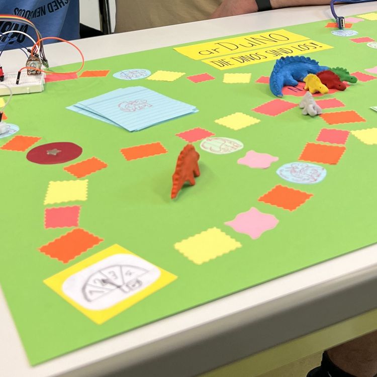 Ein Brettspiel mit grünem Spielfeld liegt auf dem Tisch, darauf bunte Felder und Dinosaurier-Spielfiguren sowie Steckplatten, aus denen Kabel ragen.