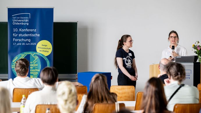Zwei junge Frauen stehen hinter und neben einem Rednerpult, links befindet sich ein Banner mit dem Logo der Konferenz. Man sieht von hinten einige Personen auf Stühlen sitzen, die den beiden zuhören. 