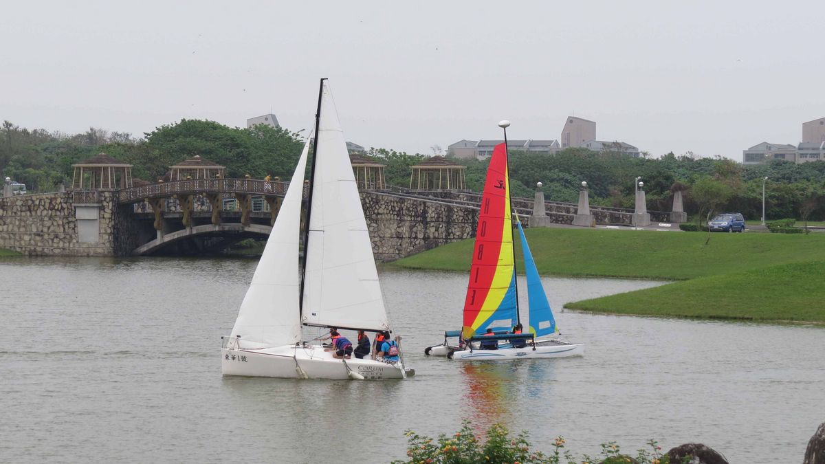 Das Bild zeigt zwei Segelboote auf einem See oder Fluss vor dem Hintergrund einer städtischen oder campusartigen Umgebung. Im Vordergrund ist ein weißes Segelboot mit mehreren Personen an Bord zu sehen, während im Hintergrund ein farbenfrohes Segelboot mit roten, blauen und gelben Segeln zu erkennen ist. Die Szene wird durch eine Brücke, üppige Grünflächen und Gebäude im Hintergrund ergänzt.