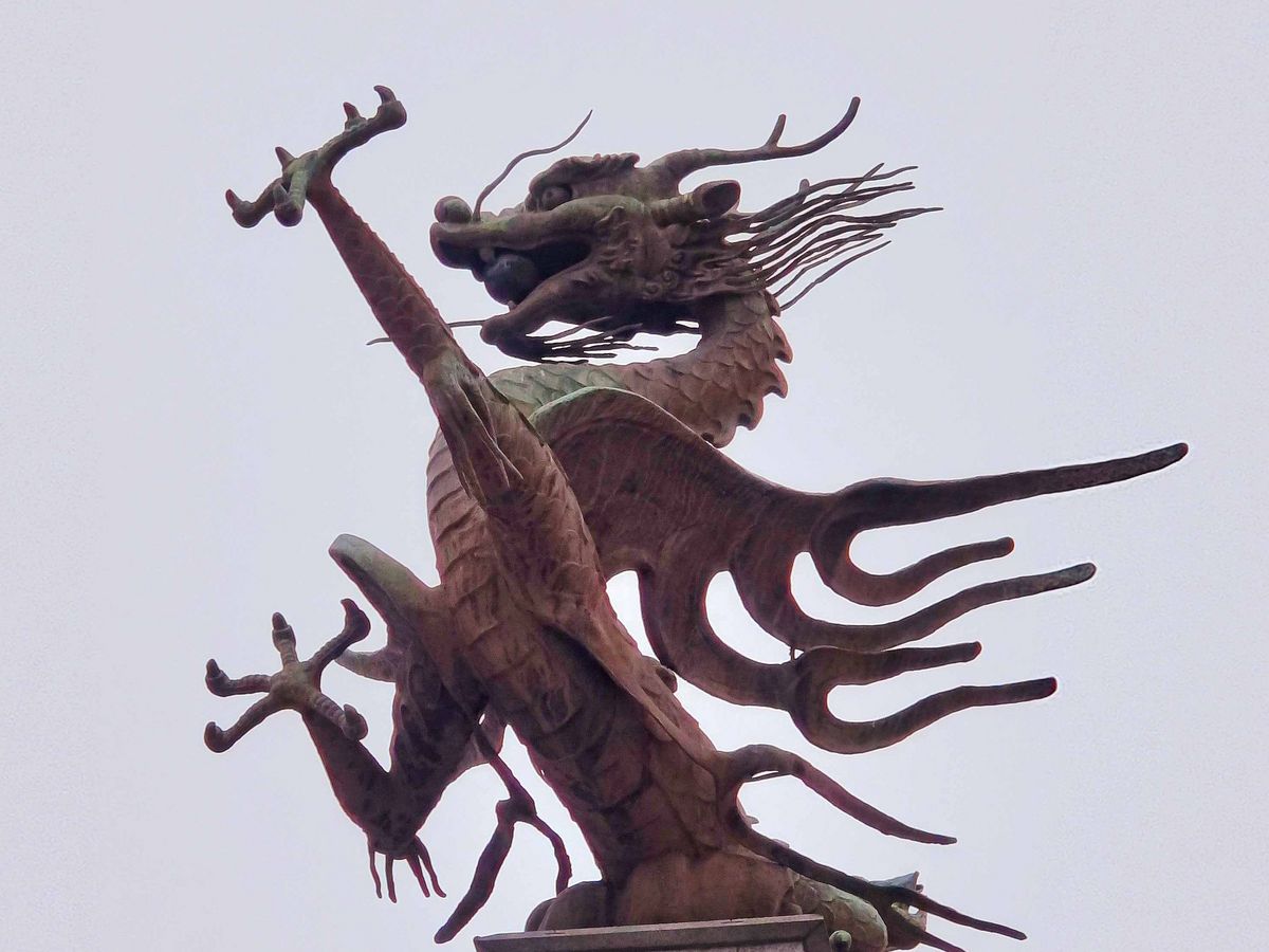 Das Bild zeigt eine große, bronzefarbene Drachenstatue, die auf einem Sockel thront. Der Drache ist in einer dynamischen Pose dargestellt, mit ausgebreiteten Flügeln und erhobenem Kopf. Der Himmel im Hintergrund ist grau und bewölkt.