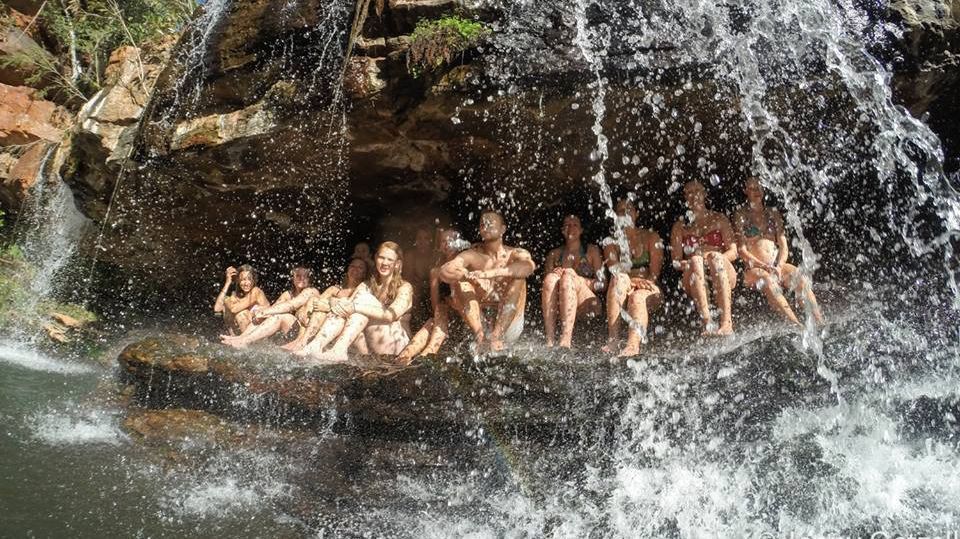 Das Bild zeigt eine Gruppe von Menschen, die hinter einem Wasserfall in einem natürlichen Becken sitzen. Das Wasser stürzt um sie herum herab und erzeugt einen sprühenden Effekt. Die Menschen scheinen die Erfahrung zu genießen und zu lachen. Die Szene wirkt erfrischend und abenteuerlich.