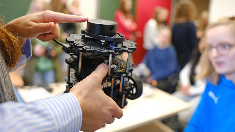 Eine Person hält eine kleine Apparatur, womöglich ein Roboter, in ihren Händen und zeigt sie einer Gruppe von Kindern.
